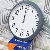 A round wall clock with a dark gray frame and white face, resting on top of a blue and orange Pixcell battery box against a tropical leaf print background.
