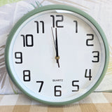A round quartz wall clock with a sage green frame and a white face, featuring large black Arabic numerals and black hands. The word 'QUARTZ' is printed below the 12.