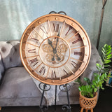 Large decorative clock with Roman numerals on a stand, placed in a room with a sofa and plant. Metal case with metal lattice and moving gears with glass face