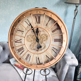 Large decorative clock with Roman numerals on a stand in a room setting. Large decorative clock with Roman numerals on a stand, placed in a room with a sofa and plant. Metal case with metal lattice and moving gears with glass face 600mm wide