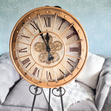 Vintage-style clock with Roman numerals on a decorative stand against a light blue textured wall. Large decorative clock with Roman numerals on a stand, placed in a room with a sofa and plant. Metal case with metal lattice and moving gears with glass face 600mm wide