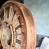 Close-up of a vintage-style clock with intricate details on a textured wall background. Large decorative clock with Roman numerals on a stand, placed in a room with a sofa and plant. Metal case with metal lattice and moving gears with glass face