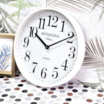 White wall clock with black numbers on a decorative surface with palm leaves and a framed picture.