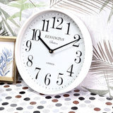 White wall clock with black numbers on a decorative surface with palm leaves and a framed picture.