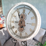 Decorative wall clock with Roman numerals on a stand against a light blue wall. met with moving gears 600mm wide