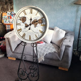 Decorative clock on a stand in a living room setting with a sofa and books. Vintage-style clock with intricate design and text highlighting features moving gears metal case glass front 600mm wide