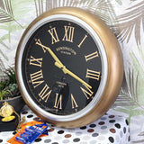 Gold-framed clock with black face and Roman numerals on a polka dot surface with plants in the background