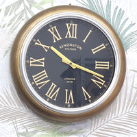 Wall clock with gold frame and black face displaying Roman numerals on a decorative background.