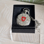 Silver pocket watch with a red emblem in a black box on a textured surface
